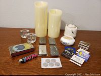 Two cream-colored flameless Luminara candles with remote controls and batteries, clear glass holders, assorted matchbooks, small wax candles, and ceramic candle holders with decorative designs shown on wooden surface.