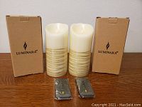 Two Luminara flameless candles with gold horizontal stripes, each with its original box and two remote controls in front.