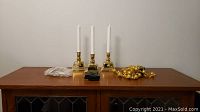 Three white Luminara taper candles in gold holders positioned on wooden cabinet with bead garlands beside them
