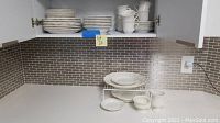 Wide view showing dinnerware plates and bowls stacked on a counter and shelves, including the small white wire shelf beneath some pieces.