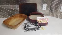 Kitchen counter setup showing stoneware sheet pan, burgundy stoneware roaster with lid, wire roaster holder, wire dish holders, and cookbook.