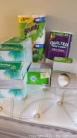 Photo showing Bounty Select-A-Size paper towels (2 rolls), 6 boxes of Kleenex tissues, an 8-roll pack of Quilted Northern Ultra Plush toilet paper, and 1 box of Puffs tissues on a mattress.
