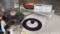 Overview showing 13-inch black Carote deep skillet, medium Revereware copper-clad pot with lid, universal glass lid with purple rim, Gotham Steel square skillet, two ice cube trays, and white wire rack on a countertop.