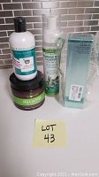 Four skincare products on white surface with tiled backsplash: Algenist boxed cleanser, Medline Remedy foaming body cleanser, Purely Northwest Tea Tree Oil body wash, Balm Shea Butter moisturizer jar.