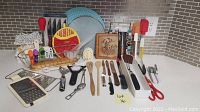 Overview of assorted kitchen utensils and accessories including scissors, spatulas, wooden spoons, graters, chip clips, trivets, bread baskets, aprons and dish towels laid out on a table.