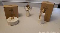 Two Willow Tree figurines standing with boxes behind, one angel with gold heart and one angel holding hands near mouth, plus a round keepsake box with angel lid.