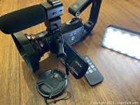 Camcorder with mounted shotgun mic, wireless mic, remote, lens cap and LED light arranged on wood surface