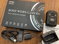 Black CamPro branded box and body worn camera with power adapter, charging dock, USB cable, instruction manual, and warranty card laid out on a wooden surface.