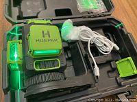 Huepar 3D green laser level inside carrying case with power cable and small green plastic accessory piece visible. Laser lines active and visible.