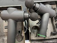 Side view of two Flyby branded massage guns in black with foam ball attachments and accessories in foam case.