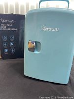 Front view of blue AstroAI portable mini refrigerator next to its box showing model and branding.