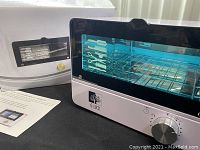 Front and side view showing the disinfection cabinet powered on with UV light glowing blue, wire racks inside, rotary timer knob, and accessories/manual placed beside it.