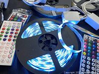 Photo showing a coiled LED strip light emitting blue light on a reel, two color remote controls, and a white control module connected to cables.