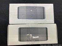 Two unopened or reclosed boxes each containing a rectangular black Bluetooth speaker from brand DOSS with packaging showing tape marks and wear.