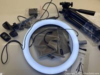 LED ring light illuminated with attached power cable, tripod stand, remote control, power adapter, and carrying bag visible.