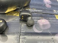 Close-up of black wireless earbud with silicone tip and charging contacts on side, placed on retail box with Zeus Air branding.