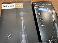 Boxes of Fairywill D7 Sonic Electric Toothbrush and Shiyog Dental Calculus Remover placed side-by-side on a wooden floor.