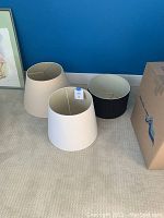 Photo showing three lampshades: a large cream-colored one with visible tear, a medium off-white one, and a smaller black one next to a cardboard box.