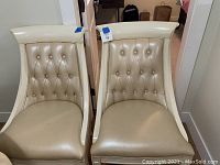 Two beige faux leather upholstered dining chairs with matching wooden frames and button tufted backs