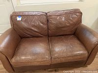 Front view of brown leather loveseat sofa showing two seat cushions and two back cushions, with some creasing and wear on the leather upholstery.