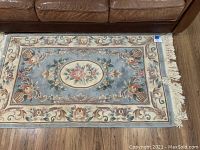 Full view of the smaller area rug on a wooden floor under a brown leather couch, showing the detailed floral pattern with blue and cream color tones and fringed edges.