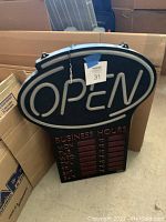 Black oval-shaped neon sign spelling 'OPEN' with attached business hours panel listing weekdays and times.