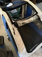 Full view of the bike attachment dog carrier showing blue and beige fabric with black mesh. Clear plastic window rolled up inside. Tear noticeable on the side fabric.