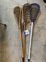 Photo showing three lacrosse sticks side by side on a concrete floor; two have wood handles and woven netting, and one has a blue plastic head with a metal handle wrapped in black tape.