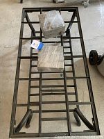 Top view of wrought iron framed table with grid layout, two stacks of square gray tiles wrapped in plastic on top.