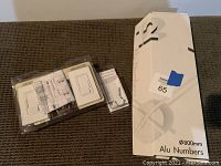 Photo showing white incandescent dimmer light switches in original packaging with instruction manuals, next to a large box labeled 'Alu Numbers' containing aluminum house numbers