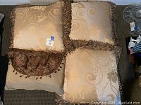 Four square pillows with beige and gold floral swirl patterns, three have gold fringe trim, one has tassel bead accent