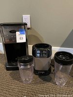 Photo shows Cuisinart coffee maker base alongside Ninja blender base and two clear blending cups with black lids.