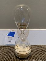Clear glass decorative hourglass with sand inside, positioned against a gray wall and carpeted floor. The hourglass includes gold accents on the upper and lower bulbs.