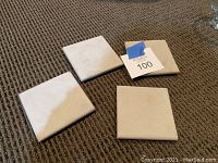 Four square stone coasters arranged on carpet, light-colored surface with visible minor discoloration and wear marks; one coaster has a blue tape with handwritten '100' attached.