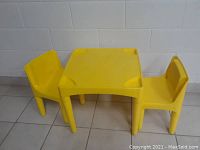 Yellow plastic children's table with two matching yellow plastic chairs placed on tiled floor against a white painted brick wall.