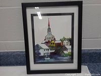 Full view of framed village scene artwork with church steeple and houses in black frame and double matting.