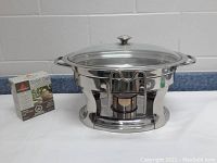 Front view of stainless steel server sitting on its stand with glass lid in place and box of solid gel fuel beside it.