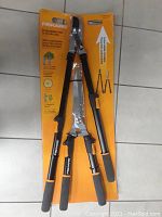 Full view of Fiskars 2-piece extendable tree and shrub trimmer set in original orange packaging, showing the tools with black handles and ergonomic grips.