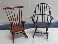 Both miniature wooden chairs placed side by side on a black and gray surface against a cinder block wall background. The rocker is natural wood tone with vertical spindles and rockers. The sack-back Windsor chair is black with natural wood accents and rounded spindle back.