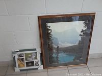 Shows large framed print with mountain and lake fishing scene next to smaller photo frame