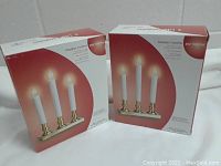 Two new in box 3-light electric candlestick chandeliers with brass plated square bases shown in original packaging.