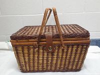 Photo shows rectangular woven basket with woven handles, leather strap with metal clasp lock at center front, and lid closed.