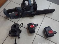 Full view of the battery operated chainsaw, two batteries and charger on floor tiles.