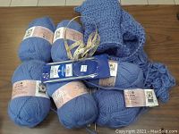 Six blue Patons Diva yarn skeins arranged on a table with a partially knitted piece, blue knitting needles in packaging, and a tape measure