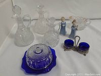 Displayed vintage glassware including three clear condiment bottles with stoppers, cobalt blue covered butter dish, cobalt blue salt cellars in silver-plated holder, two figurines, and small scalloped glass bowl.