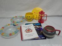 Overview of lot including colorful rimmed bowls, happy face mug and plate, French press, and books
