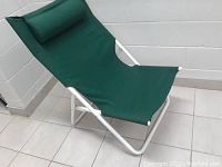 Side view of green fabric folding lounge chair on white metal frame with padded pillow, placed against white wall and tiled floor.