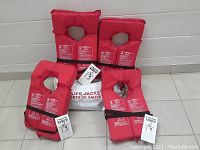 Four new, red adult personal flotation devices standing upright with white carry bag behind them, showing front labels and black straps.