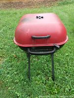 Red rectangular molded metal charcoal grill with black handle on lid and vent for airflow. Exterior and interior views showing used condition with some ash and charcoal inside.