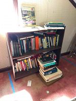 Photo showing wooden bookshelf filled with various books, some stacked on floor in front. Books cover multiple topics including religion, history, self help.